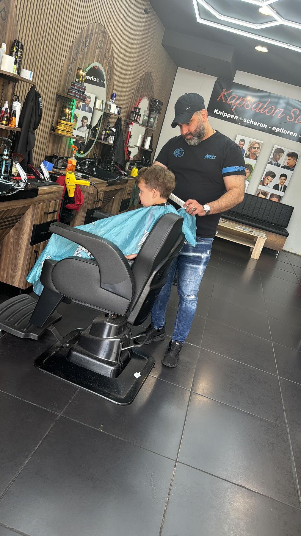 Een jongetje in de kappersstoel onder een blauwe kapmantel